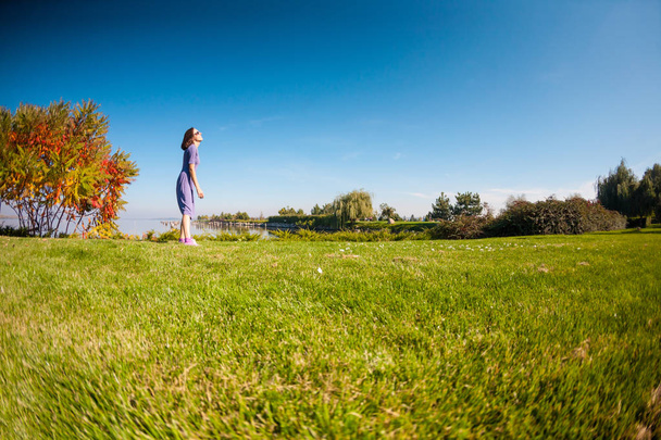 Slanke meisje wandelen langs de rivier. Een vrouw in een jurk is lopen op het groene gras. Brunette rust in het park. - Foto, afbeelding