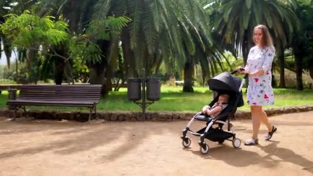 Мама прогулянки на лавці в парку з palm дерева дитини, сидячи в інвалідному візку і сміятися - Кадри, відео