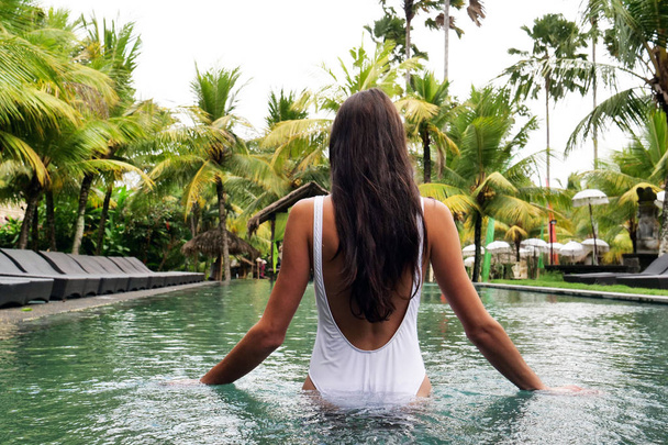 Una hermosa mujer en un resort de lujo, se relaja en las tumbonas junto a la piscina y mientras nada. Concepto de: viaje, vacaciones, relajación, hotel
. - Foto, imagen