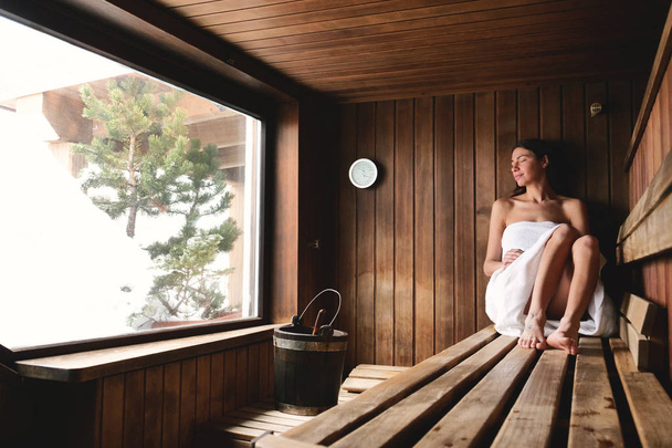 beautiful woman sitting in wooden sauna with large window - Foto, imagen