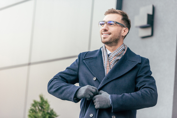 adult smiling handsome man in glasses fastening coat near building  - Fotografie, Obrázek