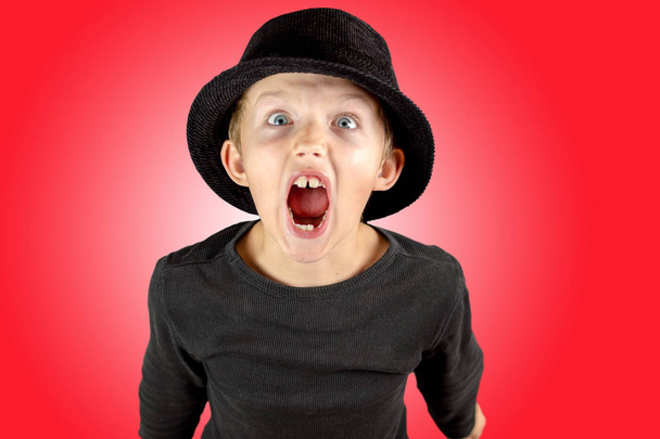 Nahaufnahme eines kleinen Jungen in schwarzem Hemd und schwarzem Hut mit schwerem schreienden Ausdruck und weit geöffneten Augen mit verrücktem Blick auf rotem Hintergrund - Foto, Bild
