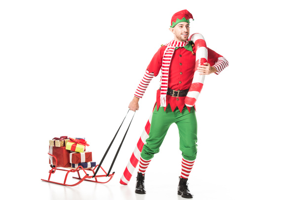 happy man in christmas elf costume carrying sleigh with presents and big candy cane isolated on white - Photo, Image