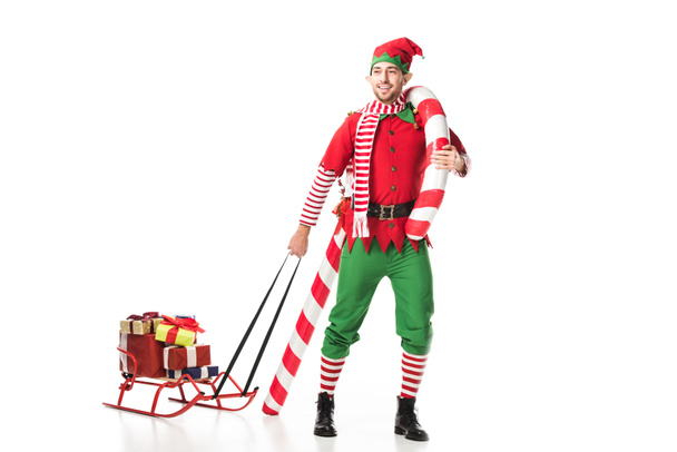 glücklicher Mann im Weihnachtselfenkostüm, der Schlitten mit Geschenken und Zuckerrohr auf weißem Grund trägt - Foto, Bild