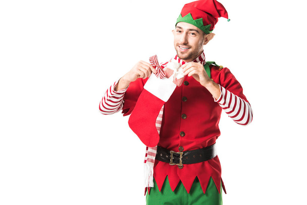 smiling man in christmas elf costume putting present in red christmas stocking and looking at camera isolated on white - Photo, Image