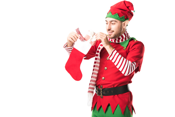 man in christmas elf costume putting present in red christmas stocking isolated on white - Photo, Image