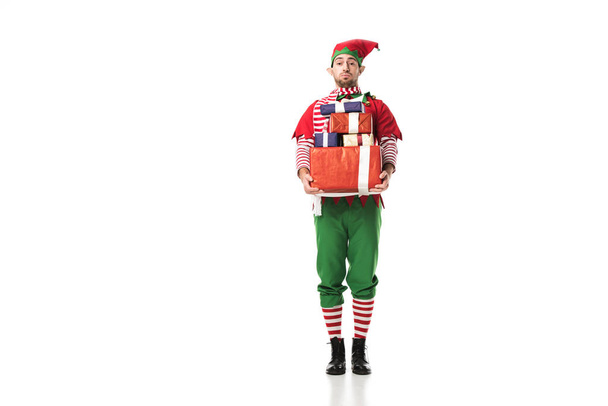 man in christmas elf costume looking at camera and carrying pile of presents isolated on white - Photo, Image