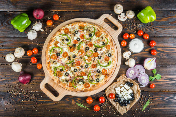 Big pizza with cheese, tomatoes, black olives and paprika on a round cutting board on a dark wooden background. Ingredients. - Foto, imagen