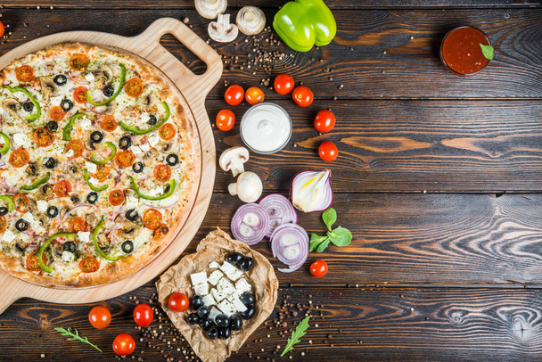 Food background design. Big pizza with cheese, tomatoes, black olives and paprika on a round cutting board on a dark wood. Ingredients. - Photo, Image