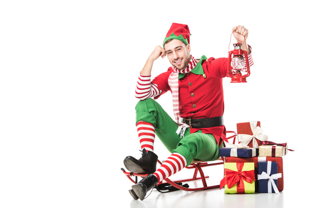 man in christmas elf costume sitting on sleigh near pile of presents and holding red lantern isolated on white - Photo, Image