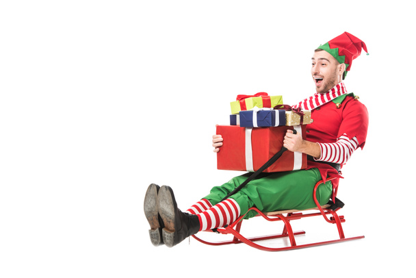 cheerful excited man in christmas elf costume holding pile of presents and riding sleigh isolated on white - 写真・画像