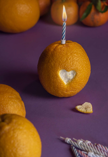 Κερί αναμμένο γενέθλια πάνω από ένα πορτοκάλι, με σχήμα καρδιάς που κόβονται από τη φλούδα, σε μωβ φόντο, περιβάλλεται από πολλά πορτοκάλια και μανταρίνια. - Φωτογραφία, εικόνα