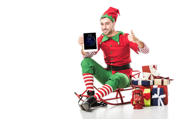 man in christmas elf costume sitting on sleigh and holding tablet with apple home screen isolated on white - Photo, image