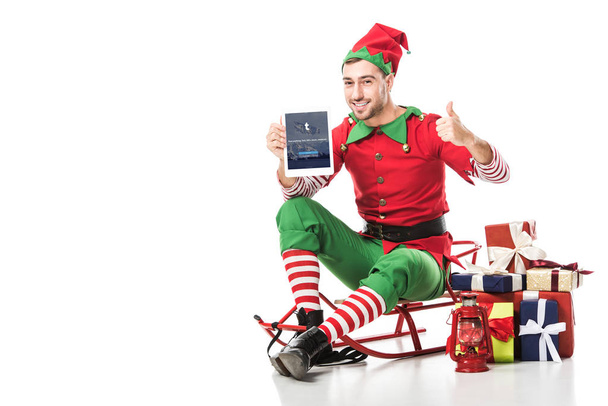 man in christmas elf costume sitting on sleigh and holding tablet with tumblr app on screen isolated on white - Photo, image