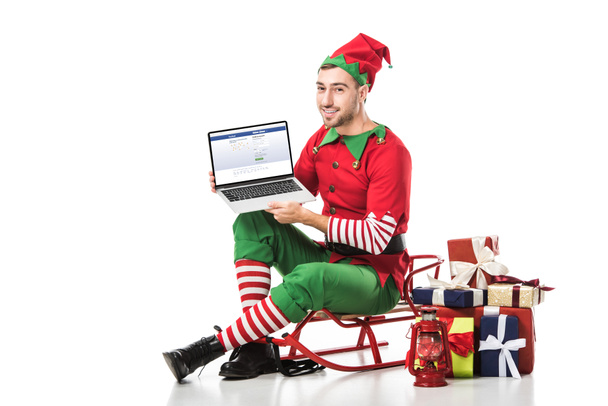 man in christmas elf costume sitting on sleigh and holding laptop with facebook website on screen isolated on white - 写真・画像