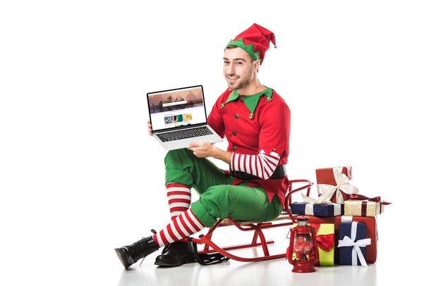 man in christmas elf costume sitting on sleigh and holding laptop with shutterstock website on screen isolated on white - Foto, Imagen