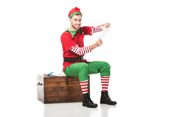 cheerful man in christmas elf costume sitting on wooden chest and holding wishlist isolated on white - Photo, Image