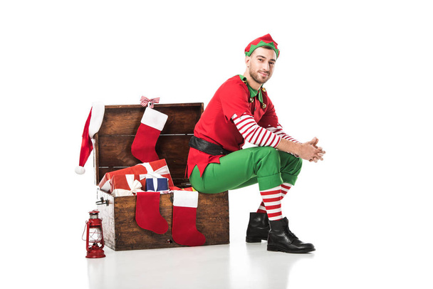 мужчина в рождественском костюме эльфа сидит на деревянном сундуке с рождественскими чулками и представляет изолированные на белом
 - Фото, изображение