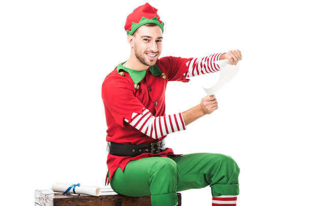 smiling man in christmas elf costume sitting and holding wishlist roll isolated on white - Photo, Image
