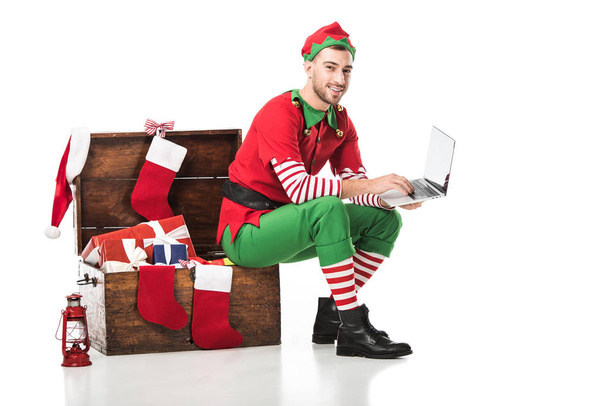 Χαμογελαστός άνθρωπος στο κοστούμι ξωτικό των Χριστουγέννων κάθεται σε ξύλινο κουτί και να χρησιμοποιούν φορητό υπολογιστή που απομονώνονται σε λευκό  - Φωτογραφία, εικόνα