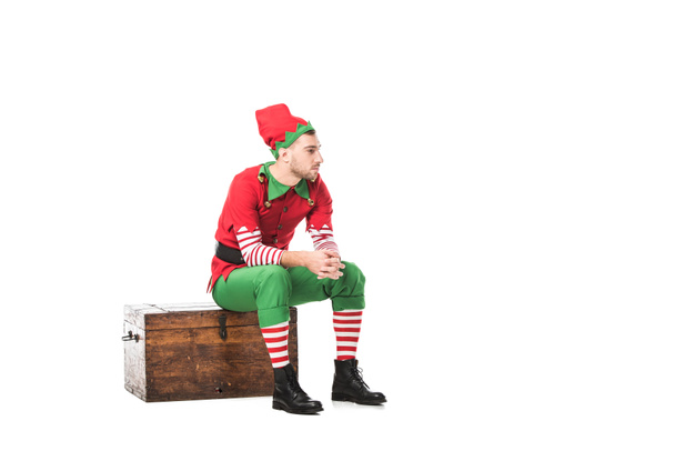pensive man in christmas elf costume sitting on wooden chest isolated on white - Photo, Image