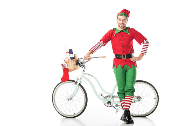 happy smiling man in christmas elf costume with hand on hip standing near bike with basket full of presents isolated on white - Valokuva, kuva