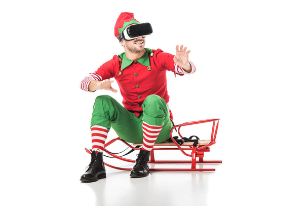 man in kerst elf kostuum zittend op een slee en dragen van virtual reality headset geïsoleerd op wit - Foto, afbeelding