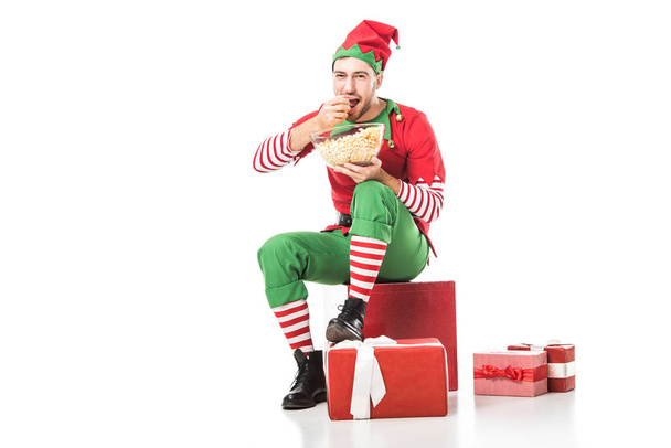 happy man in christmas elf costume sitting on pile of presents and eating popcorn isolated on white - Photo, Image