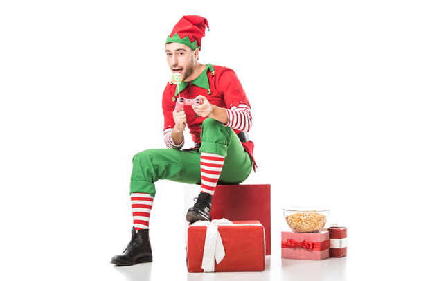 man in christmas elf costume sitting on pile of presents with lollipop and playing video game isolated on white - Photo, Image