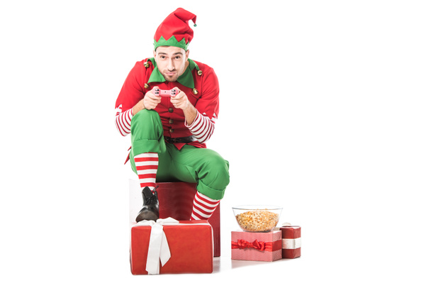 мужчина в рождественском костюме эльфа сидит на куче подарков и играет в видеоигру изолированную на белом
 - Фото, изображение