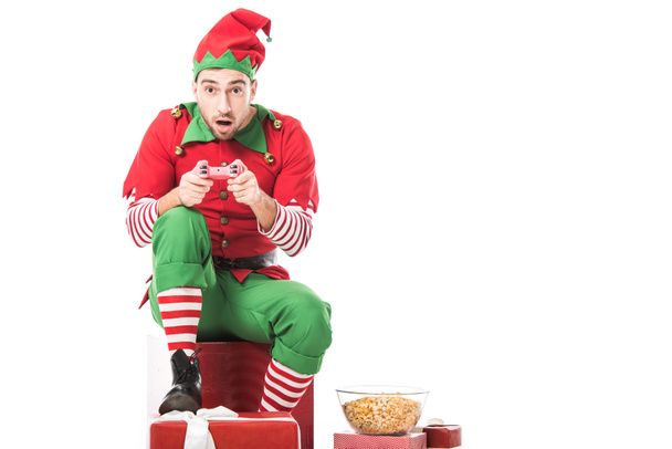 excited man in christmas elf costume sitting on pile of presents and playing video game isolated on white - Photo, Image
