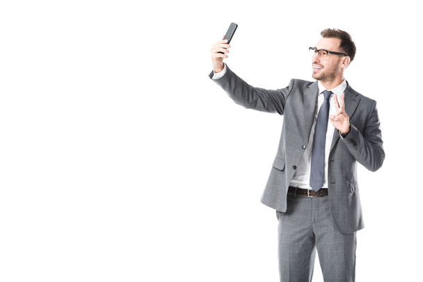 Lächelnder erwachsener Geschäftsmann macht Selfie und zeigt Friedenszeichen isoliert auf Weiß - Foto, Bild