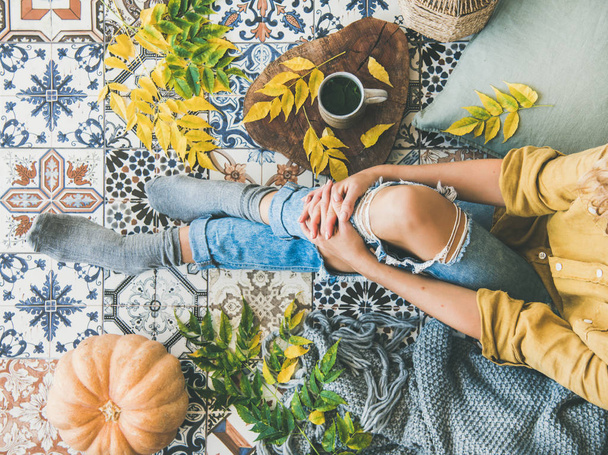 Autumn or Fall balcony tea time. Flat-lay of fallen leaves, warm blanket, pumpkin, cup of herbal tea on board and female sitting over colorful tiled floor, top view. Autumn mood concept - Фото, изображение