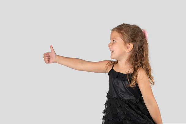 Malá usměvavá dívka, která nosí krásné černé elegantní šaty zobrazeno gesto Ok na bílém pozadí v ateliéru, pasu nahoru - Fotografie, Obrázek
