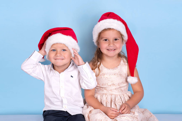 Pasa w górę portret trochę uśmiechnięte dziewczyny i chłopak sobie ubrania eleganckie i Santa Claus kapelusze. Usiąść razem i uśmiech na niebieskim tle w studio - Zdjęcie, obraz