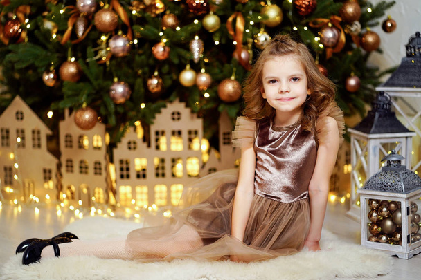 Smiling kid girl sitting under Christmas tree in room. Looking at camera. - 写真・画像