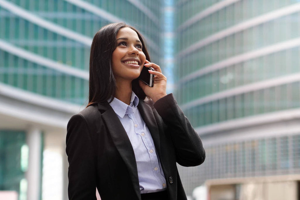 A afro-american business woman, she is happy and smiling while calling using the phone, outside the office. Concept of: technology, network, success. - Фото, зображення