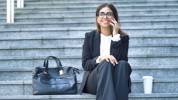 Young beautiful business woman student in suit, smiling, happy, sitting on steps, stairs, talking on the phone, coffee in plastic cup, bag, briefcase Concept new business, communication, Arab, glasses - Fotoğraf, Görsel