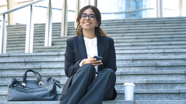 Young beautiful business woman student in suit, smiling, happy, sitting on steps, stairs, uses the phone, coffee in plastic cup, bag, briefcase Concept new business, communication, Arab, glasses - Photo, image