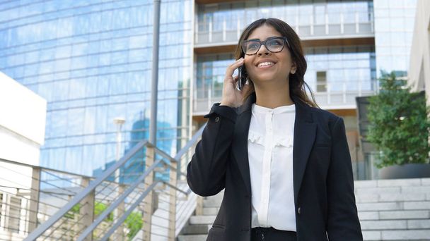 Young beautiful business woman (student) in a suit, smiling, happy, going down stairs, steps, talking on the phone, writing a message. Concept: new business, communication, Arab, banker, glasses, call - Photo, image