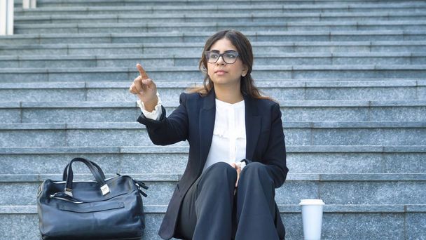 Arab business woman touching with finger futuristic invisible virtual hologram and sitting on stairs outdoors  - Foto, Imagen