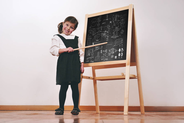 A happy little girl dressed as a teacher in front of a small blackboard holds a lesson in economics, marketing, teamwork, mathematics. Concept of: educational, school, business and love for study. - Фото, зображення