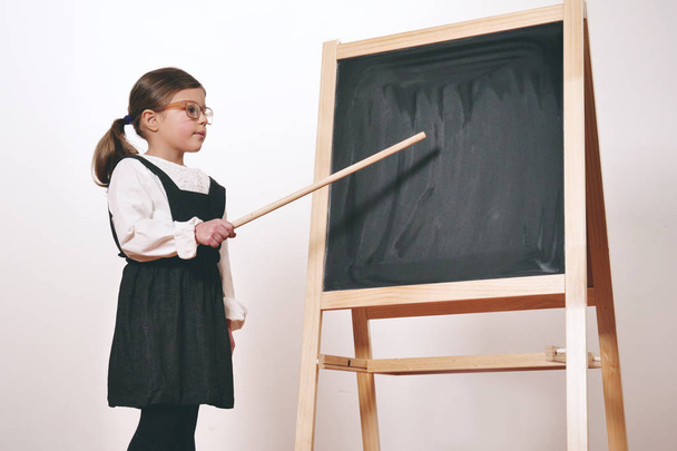 A happy little girl dressed as a teacher in front of a small blackboard holds a lesson in economics, marketing, teamwork, mathematics. Concept of: educational, school, business and love for study. - Photo, Image