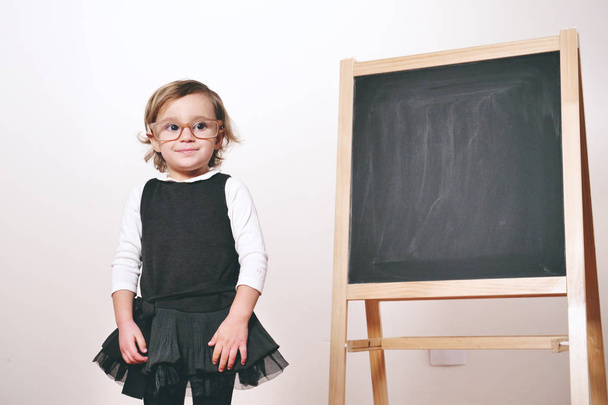 A happy little girl dressed as a teacher in front of a small blackboard holds a lesson in economics, marketing, teamwork, mathematics. Concept of: educational, school, business and love for study. - Valokuva, kuva