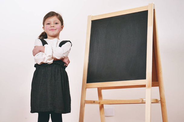 A happy little girl dressed as a teacher in front of a small blackboard holds a lesson in economics, marketing, teamwork, mathematics. Concept of: educational, school, business and love for study. - Photo, Image