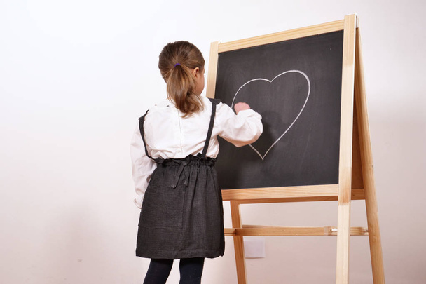 Portrait of a happy little girl dressed as a teacher in front of a small blackboard, she draws a heart with chalk as a sign of love for school. Concept of: educational, school, business - Photo, Image