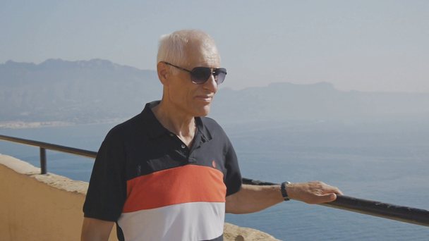 Bejaarde man schakelde toeristische. Kaukasische grijs-haired senior man in zonnebril lopen op hoge verhoogde observatie dek op zee kust achtergrond. - Video
