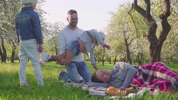 Šťastná rodina tráví čas na piknik poblíž jabloni. Matka leží na trávě, děti a manžela k ní. Každý líbá matka. - Záběry, video