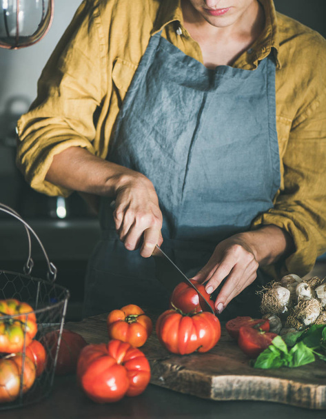Woman in grey linen apron cutting and cooking tomato sauce, canned tomatoes or pasta with heirloom tomatoes, basil and garlic at kitchen counter. Seasonal healthy cooking, slow food or comfort food - Foto, Imagen
