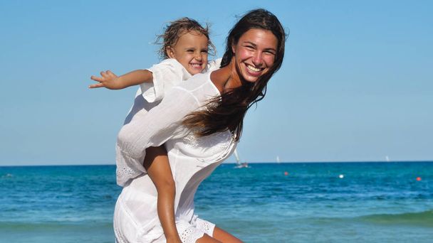 Mother and daughter happily playing at sea, hugging in white clothes smiling, background of sea blue water. - Фото, зображення